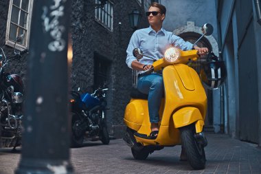 Genç adam şık giyinmiş bir beyaz bir gömlek ve pantolon akşam eski bir Europe street üzerinde sarı klasik İtalyan scooter üzerinde binmek.