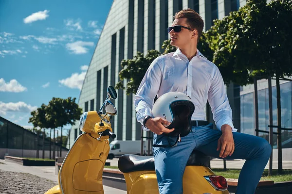 Emin yakışıklı şık saç kesimi ile beyaz bir gömlek, kot pantolon ve güneş gözlüğü tutar kask ve bir gökdelen karşı sarı klasik İtalyan scooter üzerinde oturan giymiş.