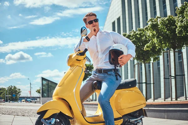 Dalgın yakışıklı adam beyaz bir gömlek, kot pantolon ve güneş gözlüğü giymiş şık saç kesimi ile kask ve el tutar çeneme bir gökdelen karşı sarı klasik İtalyan scooter üzerinde otururken.