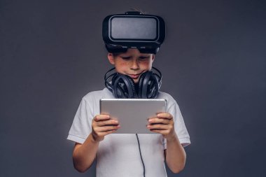 Küçük bir çocuk portresi olan sanal gerçeklik gözlük ve kulaklık beyaz bir tişört giymiş ve gri arka plan üzerinde izole bir stüdyoya dijital tablet kullanma. Eğitim ve teknoloji kavramı.