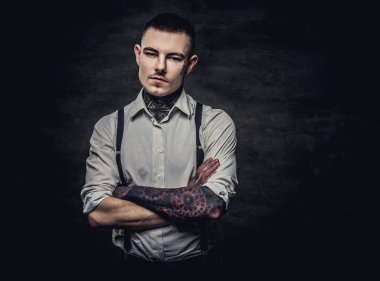 Genç eski moda dövmeli adam giyen beyaz gömlek ve pantolon askısı, poz ile çapraz kollar, bir kameraya bakıyor. Karanlık bir arka plan üzerinde izole.