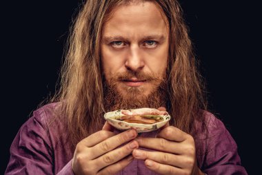 Yakın çekim portre bir mutlu kızıl saçlı hippi erkek uzun saç ve tam sakal mor bir gömlek giymiş bir kameraya bakarak çay ile küçük bir daire tutar. Karanlık dokulu bir arka plan üzerinde izole.