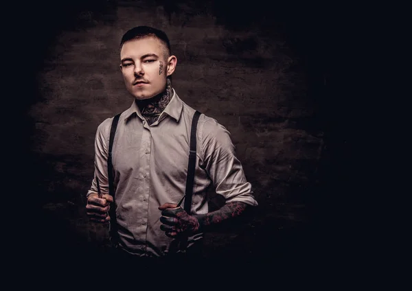 Portrait of a old-fashioned tattooed hairdresser wearing a white shirt with suspenders holds a scissors. Isolated on a dark textured background.