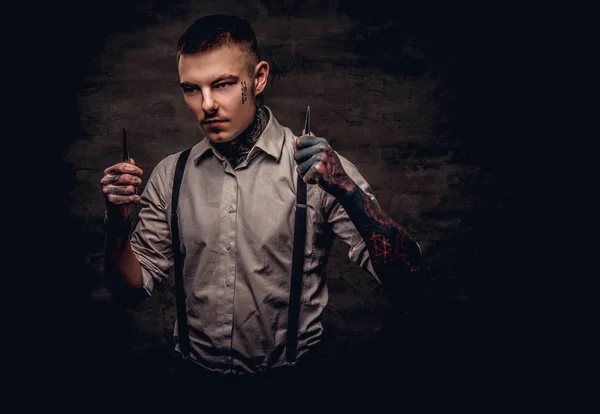 Portrait of a old-fashioned tattooed hairdresser wearing a white shirt with suspenders holds a scissors. Isolated on a dark textured background.