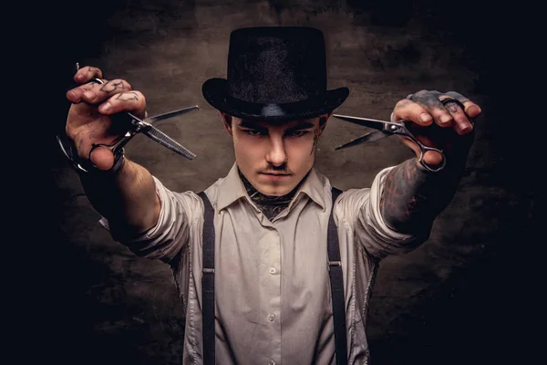 Portrait of a old-fashioned tattooed hairdresser wearing a white shirt with suspenders and cylinder hat, looking at a camera, holds a scissors. Isolated on a dark textured background.