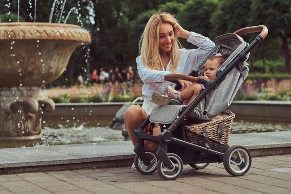 幸福的金发女郎穿着白色上衣和短裤 她的小女儿在一个婴儿推车附近的喷泉在城市公园 — 图库照片