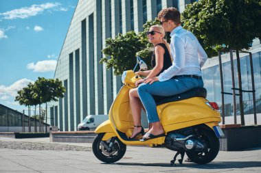 Güzel çift - şık yakışıklı olan beyaz bir tişört ve kot pantolon ve sevimli sarışın kadın birlikte bir gökdelen karşı sarı bir retro scooter üzerinde oturan siyah elbise giymiş. Yaz Avrupa tatil, tarih, romantizm.