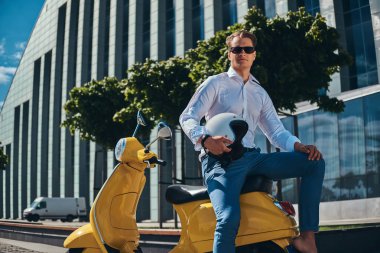 Emin yakışıklı şık saç kesimi ile beyaz bir gömlek, kot pantolon ve güneş gözlüğü tutar kask ve bir gökdelen karşı sarı klasik İtalyan scooter üzerinde oturan giymiş.