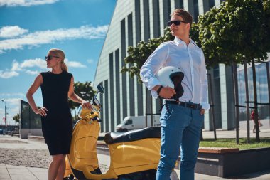 Beyaz tişört ve kot pantolon ve siyah elbise ayakta bir sarı klasik İtalyan scooter yakınındaki bir gökdelen karşı giyen sevimli sarışın kadın çekici çift - yakışıklı olan şık giyinmiş. Tatil ve insanlar kavramı.
