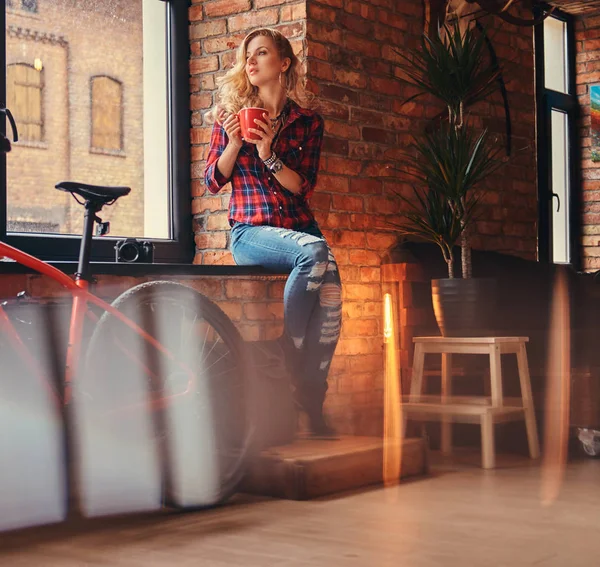 Uzun kıvırcık saçlı dalgın şehvetli Sarışın hipster kadın bir polar gömlek giymiş ve kot tutar uzağa arıyorsunuz bir pencere pervazına üzerinde bir çatı katı iç bir stüdyoda oturup sabah kahve.