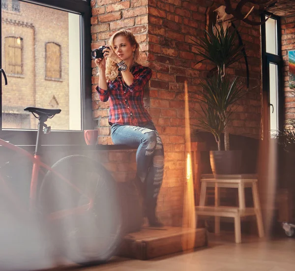 Uzun kıvırcık saçlı kızla şehvetli Sarışın hipster bir polar gömlek giymiş ve kot tutan bir pencere pervazına çatı iç bir stüdyoda oturup bir kamera.