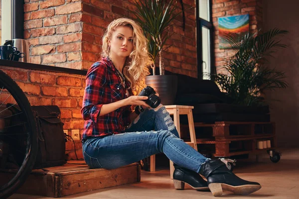 Sensual menina hipster loira com cabelos longos encaracolados vestida com uma camisa de lã e jeans mantém uma câmera sentada em uma caixa de madeira em um estúdio com interior loft . — Fotografia de Stock
