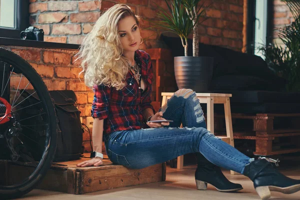 Sensual chica hipster rubia con el pelo largo rizado vestido con una camisa de lana y jeans sostiene un teléfono inteligente sentado en una caja de madera en un estudio con un loft interior . —  Fotos de Stock