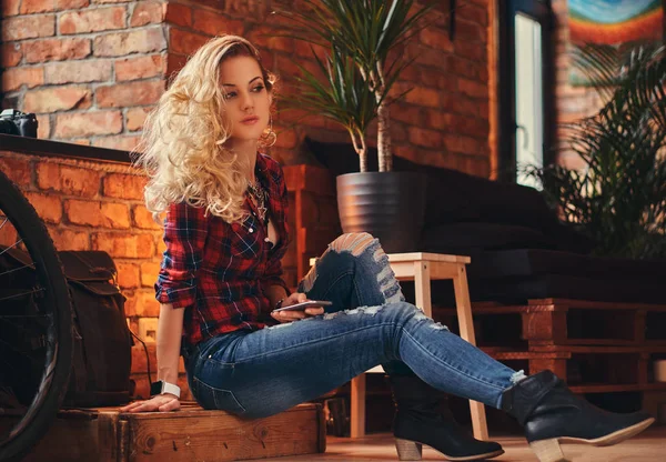 Smyslné blond bokovky dívka s dlouhými kudrnatými vlasy oblečený v fleecová košile a džíny drží smartphone seděl na dřevěné krabici ve studiu s interiér podkroví. — Stock fotografie