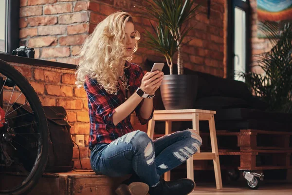 Sensuele blonde hipster meisje met lang krullend haar gekleed in een fleece shirt en spijkerbroek houdt een smartphone zittend op een houten kist in een studio met een loft interieur. — Stockfoto