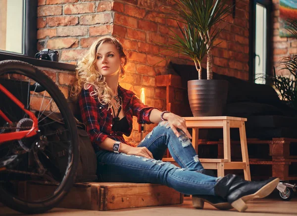 Sensual blonde hipster girl with long curly hair dressed in a fleece shirt and jeans sitting on a wooden box, looking away, at a studio with loft interior. — Stock Photo, Image