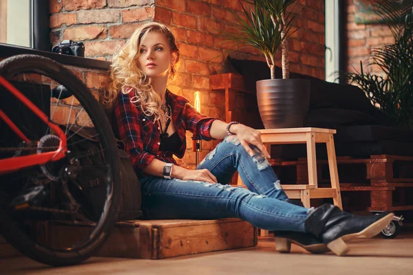 Sensuell blond hipster flicka med långt lockigt hår iklädd en fleece skjorta och jeans som sitter på en trälåda, tittar bort, vid en studio med loft interiör. — Stockfoto