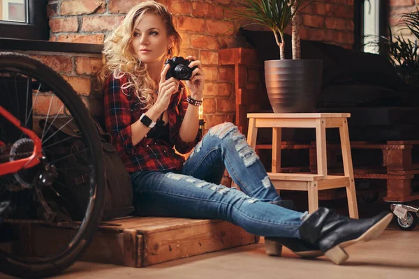 Sensuell blond hipster flicka med långt lockigt hår klädd i en fleece skjorta och jeans håller en kamera som sitter på en trälåda på en studio med loft interiör. — Stockfoto
