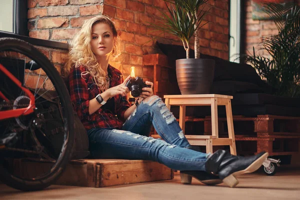 Sensual chica hipster rubia con el pelo largo rizado vestido con una camisa polar y jeans sostiene una cámara sentada en una caja de madera en un estudio con interior loft . —  Fotos de Stock
