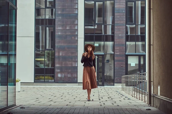 Moda donna elegante indossando una giacca nera, cappello marrone e gonna con una pochette borsetta passeggiando sul centro città europeo . — Foto Stock