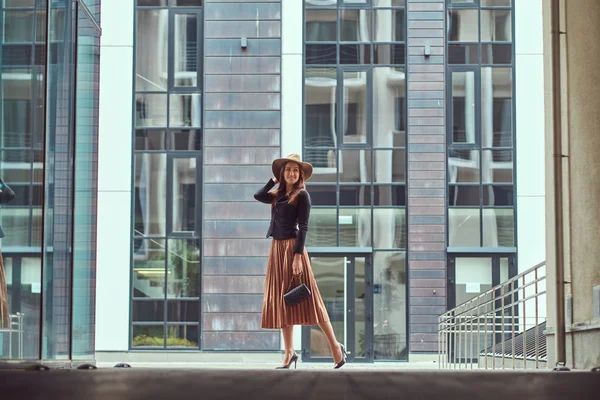 Moda feliz mulher elegante vestindo uma jaqueta preta, chapéu marrom e saia com uma embreagem bolsa andando no centro da cidade europeia . — Fotografia de Stock