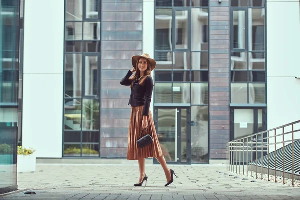 Mujer elegante moda feliz con una chaqueta negra, sombrero marrón y falda con un bolso de embrague caminando por el centro de la ciudad europea . —  Fotos de Stock