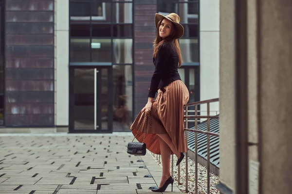 Mujer elegante de moda sonriente con una chaqueta negra, sombrero marrón y falda con un bolso de mano posando mientras se apoya en una barandilla de acero en el centro de la ciudad europea . — Foto de Stock