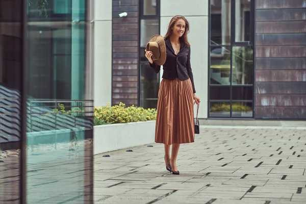 Full body portrait of a happy fashion elegant woman wearing a black jacket and skirt holds a hat and handbag clutch walking on a European city center.