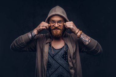 Gülümseyen Kızıl saçlı hippi tam sakal ve gözlük bir stüdyoda hoodie ve t-shirt pozlar giymiş. Koyu arka plan üzerinde izole.