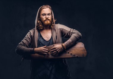 Redhead hipster male with long luxuriant hair and full beard dressed in a hoodie and t-shirt playing on a Russian traditional musical instrument - gusli. Isolated on the dark background.  clipart