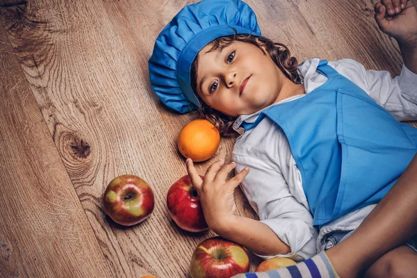 Bir mavi aşçı Tekdüzen bir ahşap zemin üzerinde meyve ile yalan giyinmiş kahverengi kıvırcık saçlı küçük çocuk.