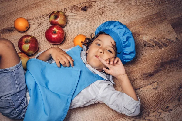 Bir mavi aşçı Tekdüzen bir ahşap zemin üzerinde meyve ile yalan giyinmiş kahverengi kıvırcık saçlı küçük çocuk.