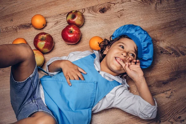 Bir mavi aşçı Tekdüzen bir ahşap zemin üzerinde meyve ile yalan giyinmiş kahverengi kıvırcık saçlı küçük çocuk.