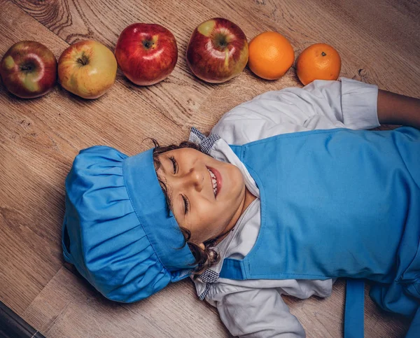 Bir mavi aşçı Tekdüzen bir ahşap zemin üzerinde meyve ile yalan giyinmiş kahverengi kıvırcık saçlı küçük çocuk.
