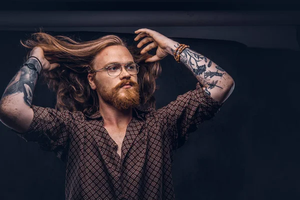 Tattoed Kızıl saçlı adam hipster gür saçları bir stüdyoda ayakta bir kahverengi gömlek giymiş düzeltir. Koyu arka plan üzerinde izole.