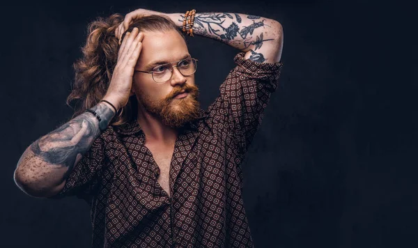 Tattoed redhead man hipster corrects his lush hair dressed in a brown shirt, standing at a studio. Isolated on the dark background.