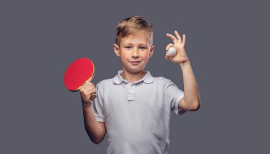 Kızıl saçlı okullu çocuk beyaz bir t-shirt giymiş bir stüdyoda bir ping-pong raket ve top tutar. Gri arka plan üzerinde izole.