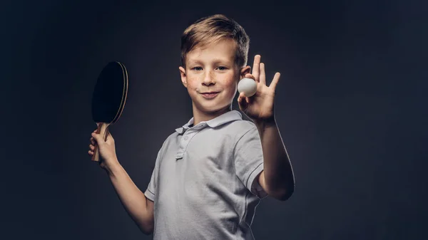 身穿白色 T恤衫的红发小学生在画室里拿着乒乓球球拍和球 在灰色背景上隔离 — 图库照片