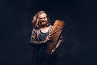 Gülümseyen Kızıl saçlı hippi erkek uzun gür saçlı ve tam sakal içinde rahat kıyafetler giymiş bir Rus geleneksel müzik aleti - gusli tutar. Karanlık bir arka plan üzerinde izole.