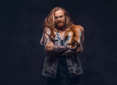 Tattoed Kızıl saçlı hippi erkek uzun gür saçlı ve tam sakal bir t-shirt giymiş ve bir stüdyoda bir omuz fox deri ceket tutar. Karanlık bir arka plan üzerinde izole.