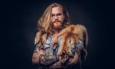 Tattoed Kızıl saçlı hippi erkek uzun gür saçlı ve tam sakal bir t-shirt giymiş ve bir stüdyoda bir omuz fox deri ceket tutar. Karanlık bir arka plan üzerinde izole.