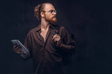 Gözlüklü bir kahverengi gömlek giymiş bir kızıl saçlı hipster öğrenci portresi tutan bir sırt çantası ve bir stüdyoda poz dijital tablet. Koyu arka plan üzerinde izole.