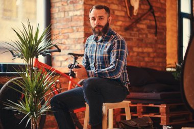 Yakışıklı sakallı hippi erkek mavi Polar gömlek ve pantolon bir kamera bir çatı katı iç ile bir stüdyo ahşap Stool'da üzerinde otururken tutan bir kameraya bakıyor.