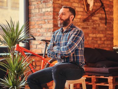 Yakışıklı sakallı hippi erkek mavi Polar gömlek ve bir çatı katı iç ile bir stüdyo ahşap Stool'da çapraz silah ile oturan kot.