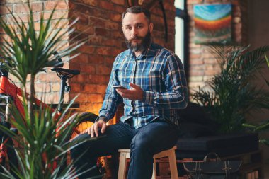 Studio loft iç ahşap bir dışkı üzerinde otururken bir smartphone yakışıklı sakallı hippi bir mavi Polar gömlek ve kot pantolon erkek tutar.