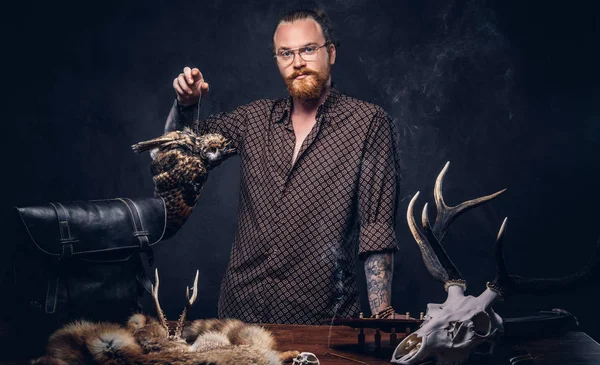 Kızıl saçlı kesişiyor hippi erkek güneş gözlüğü el yapımı kupa, baykuş korkuluk ve fox deri bir tabloyla yakın duran bir kahverengi gömlek giymiş. Karanlık bir arka plan üzerinde izole.