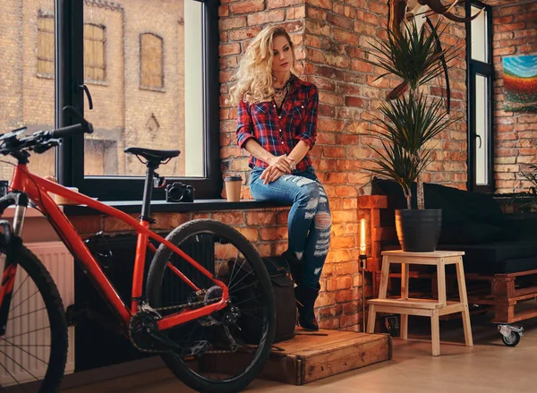 Uzun kıvırcık saçlı kızla şehvetli Sarışın hipster bir polar gömlek giymiş ve kot tutan bir pencere pervazına çatı iç bir stüdyoda oturup sabah kahve.