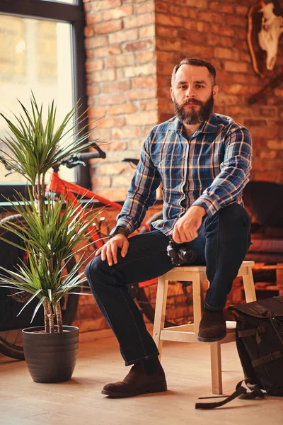 Bir kameraya bakarak bir çatı katı iç ile bir stüdyo ahşap Stool'da üzerinde otururken yakışıklı sakallı hippi bir mavi Polar gömlek ve kot pantolon erkek portresi bir kamera tutan.