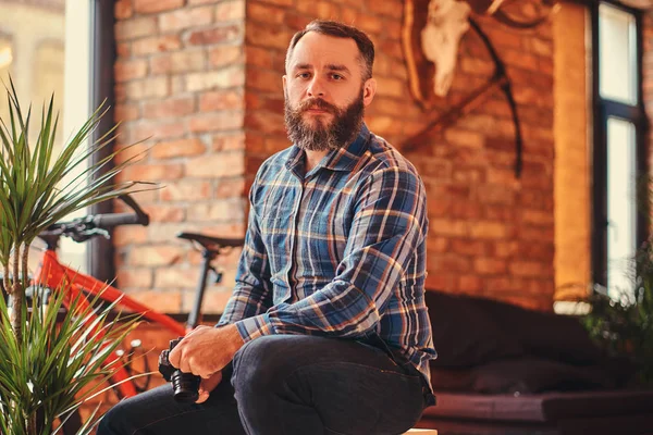 Bir kameraya bakarak bir çatı katı iç ile bir stüdyo ahşap Stool'da üzerinde otururken yakışıklı sakallı hippi bir mavi Polar gömlek ve kot pantolon erkek portresi bir kamera tutan.