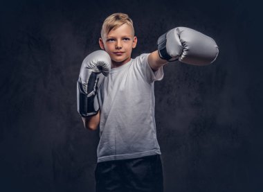 Okul çocuğu boksör sarı saçlı bir beyaz t-shirt giyen boks eldiven giymiş bir boks kick gösterir. Karanlık dokulu arka plan üzerinde izole.